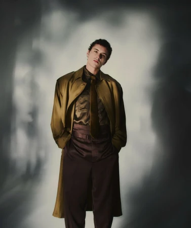 Stylish young man in olive trench coat, brown shirt and pants, standing in studio with soft lighting and diffused background.