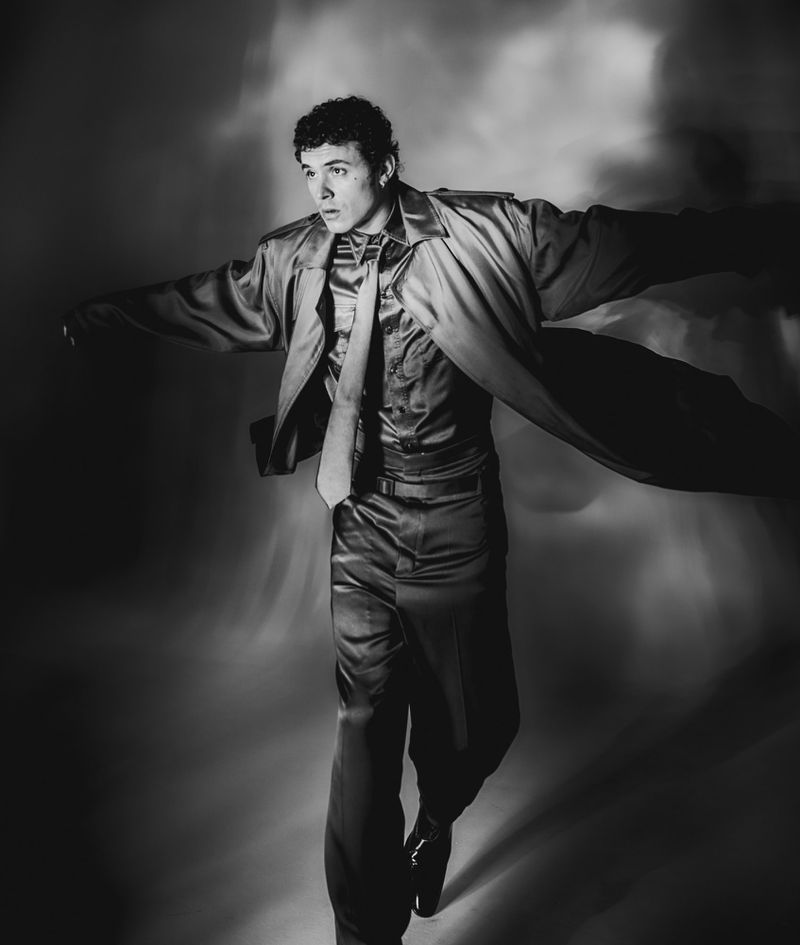 Monochromatic portrait of a young man in a shiny suit and tie with an open coat, posing in a neutral studio backdrop with diffuse lighting.