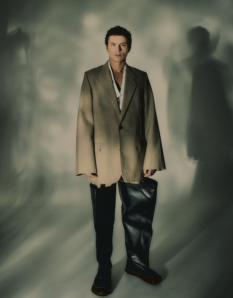 A person wearing an oversized blazer, black pants, and a single tall leather boot, standing in a photo studio against a mottled background.
