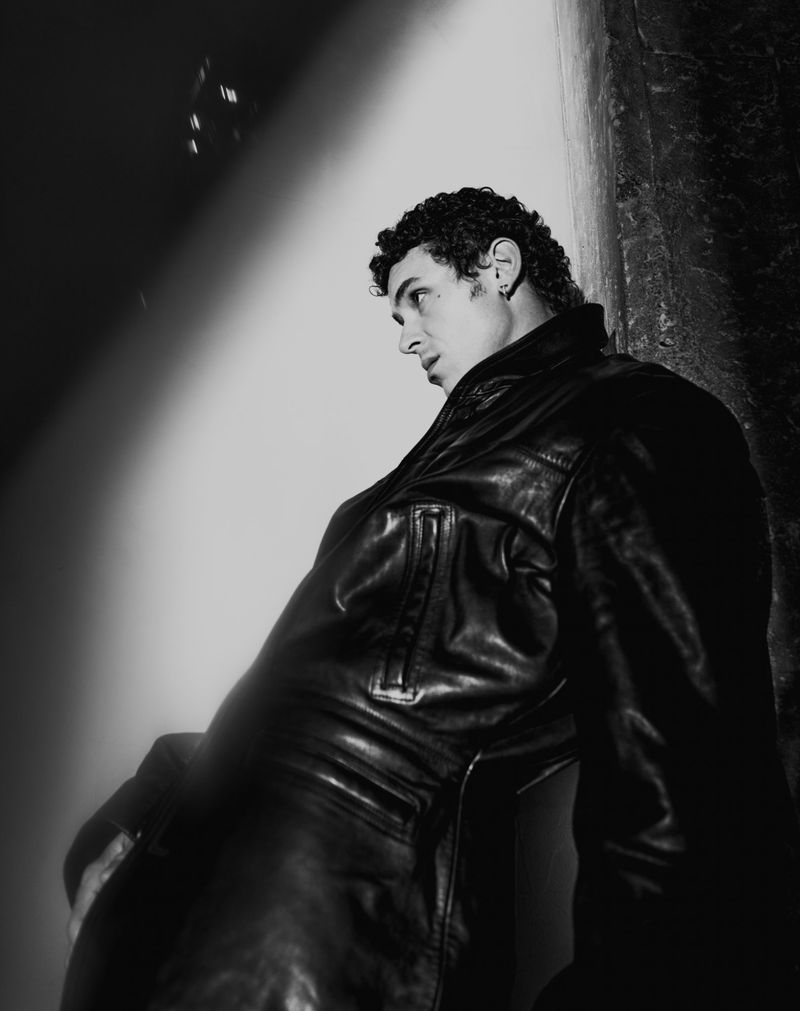 Black and white shot of a young adult male in a leather jacket leaning against a wall, gazing off to the side with a serious expression.