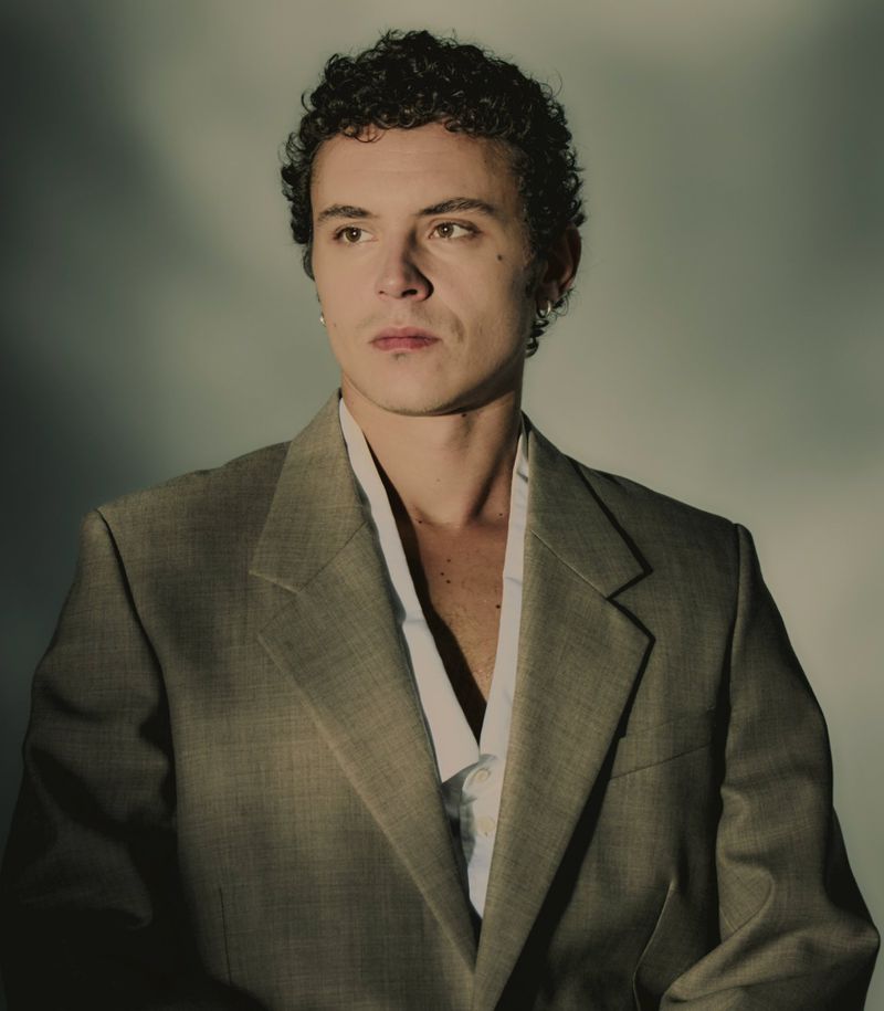 Portrait of a young adult male with curly hair wearing a gray suit jacket, looking slightly off-camera against a neutral background.