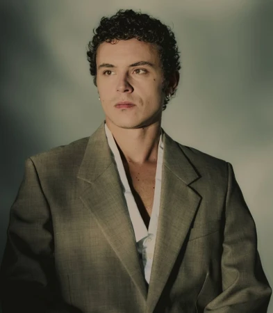 Portrait of a young adult male with curly hair wearing a gray suit jacket, looking slightly off-camera against a neutral background.