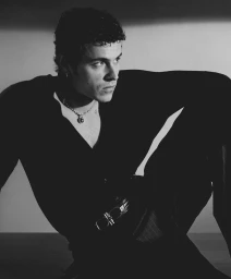 Portrait of actor Aron Piper wearing a dark shirt and chain necklace against a light background.