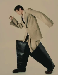 A man wearing an oversized beige suit jacket and exaggerated black boots running against a neutral backdrop
