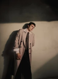 Portrait of Aron Piper wearing a beige double-breasted trench coat and striped tie against a plain wall.