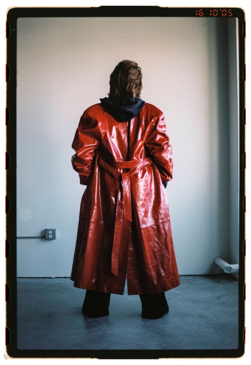 Back view of a person in a red vinyl trench coat with a hoodie, standing against a plain wall in an empty room with a concrete floor.