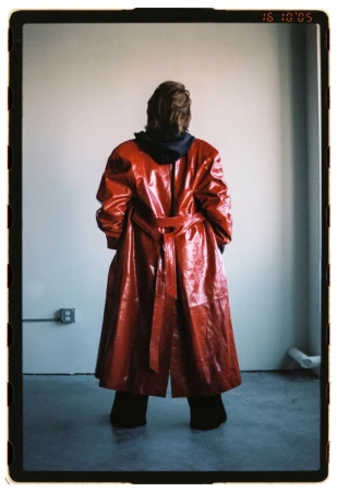 Back view of a person in a red vinyl trench coat with a hoodie, standing against a plain wall in an empty room with a concrete floor.
