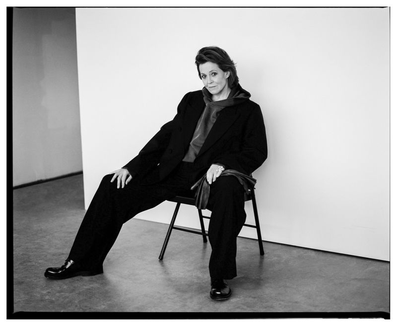 Portrait of Sigourney Weaver seated on a chair in a dark suit jacket and trousers against a white studio wall