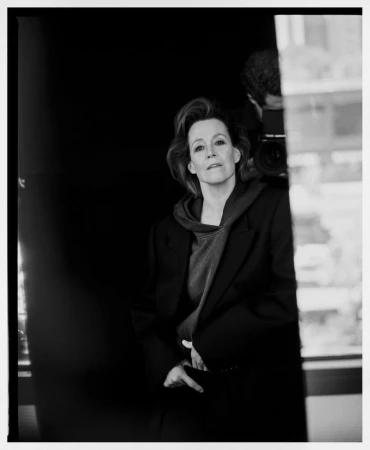 Black and white portrait of Sigourney Weaver in a dark tailored jacket with a photographer holding a camera visible in reflection.