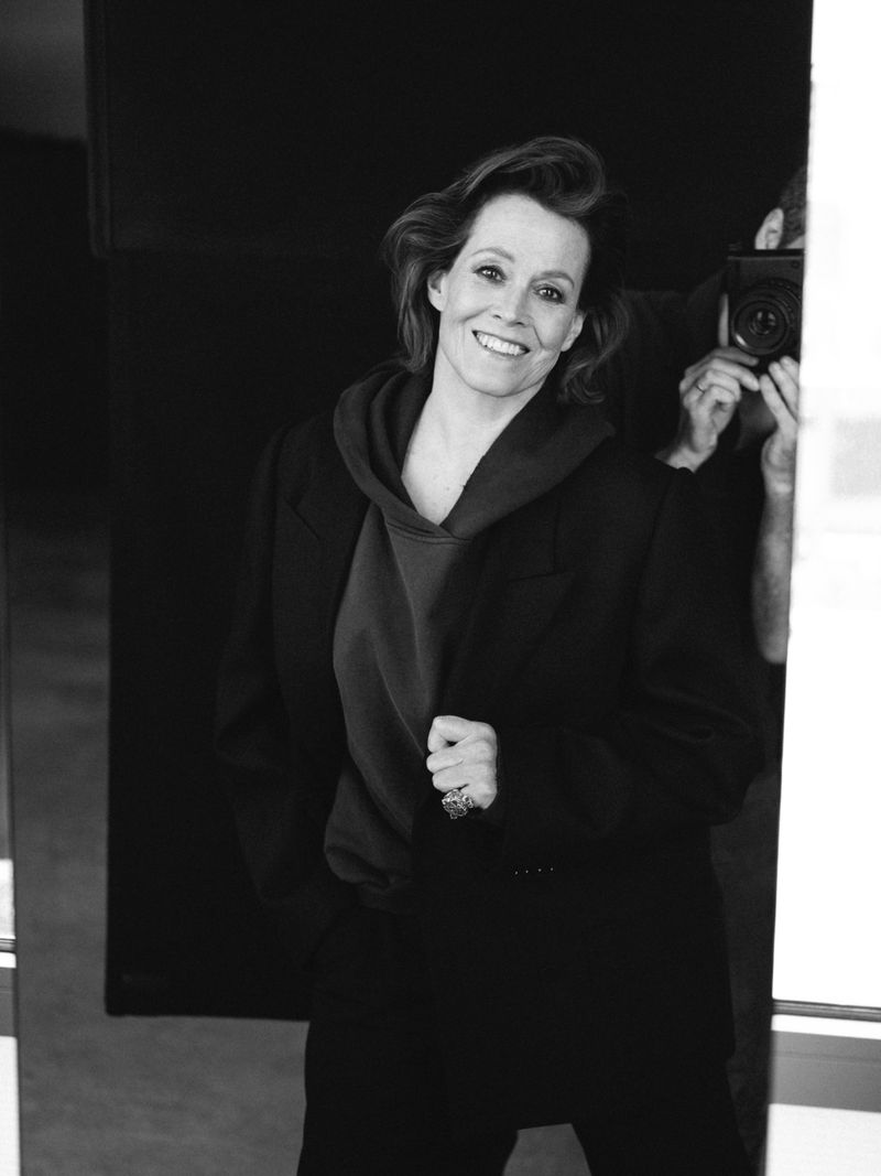 Black-and-white portrait of Sigourney Weaver indoors, wearing a dark jacket and hoodie. A photographer is visible in the background.