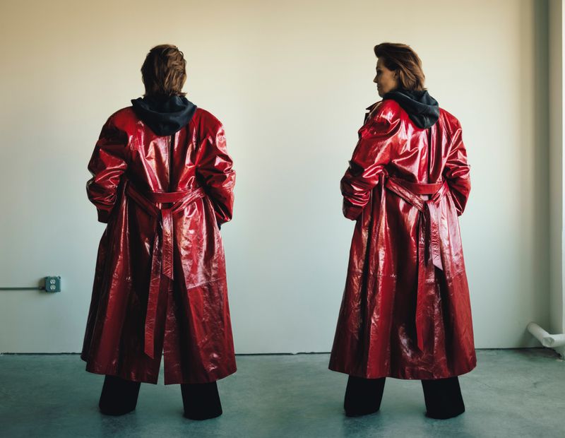 Two figures wearing shiny red belted coats with black hoodies stand against a neutral wall, one looking back at the viewer.