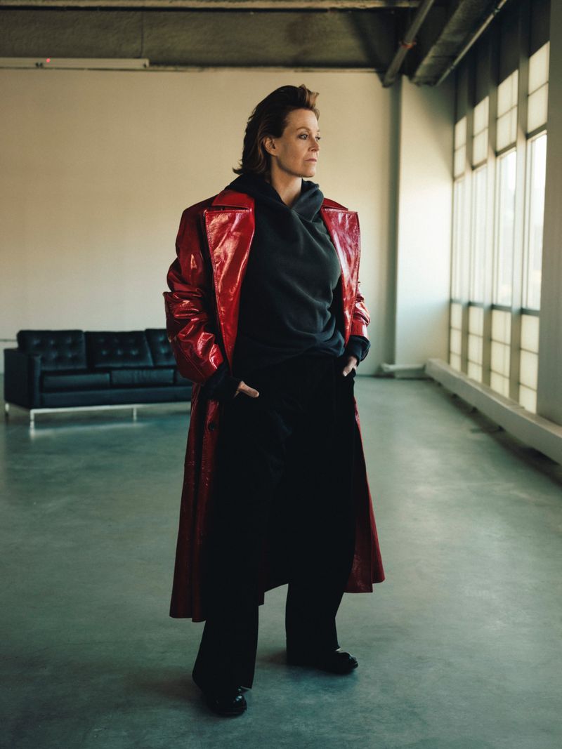 Indoor studio shot of an adult woman in a red patent leather coat and black outfit standing against a neutral wall with a sofa.
