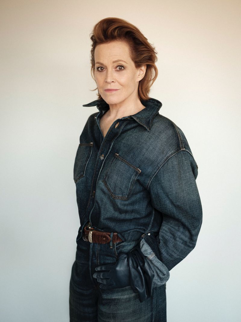 Portrait of Sigourney Weaver in denim shirt and pants, hands in black gloves, posed in front of a bright neutral studio backdrop.