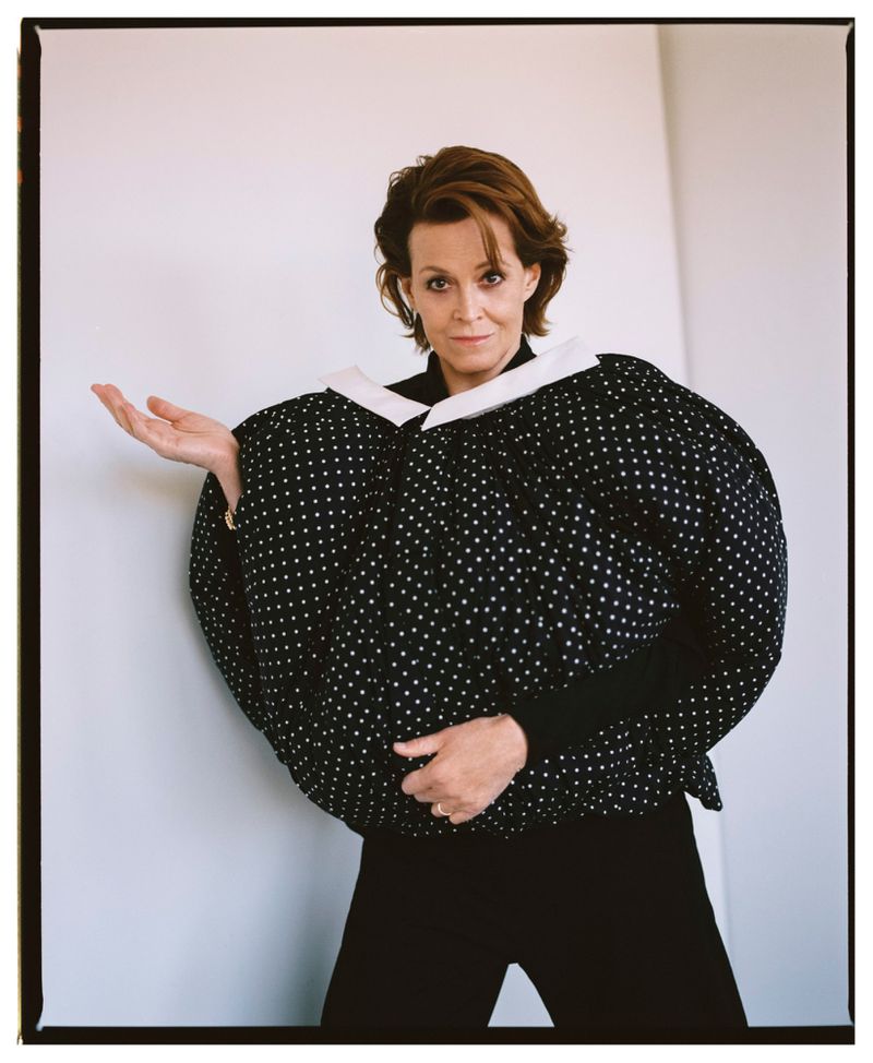 Elegant woman in a black-and-white polka dot jacket poses indoors against a white wall with open palm, in a medium-shot portrait.