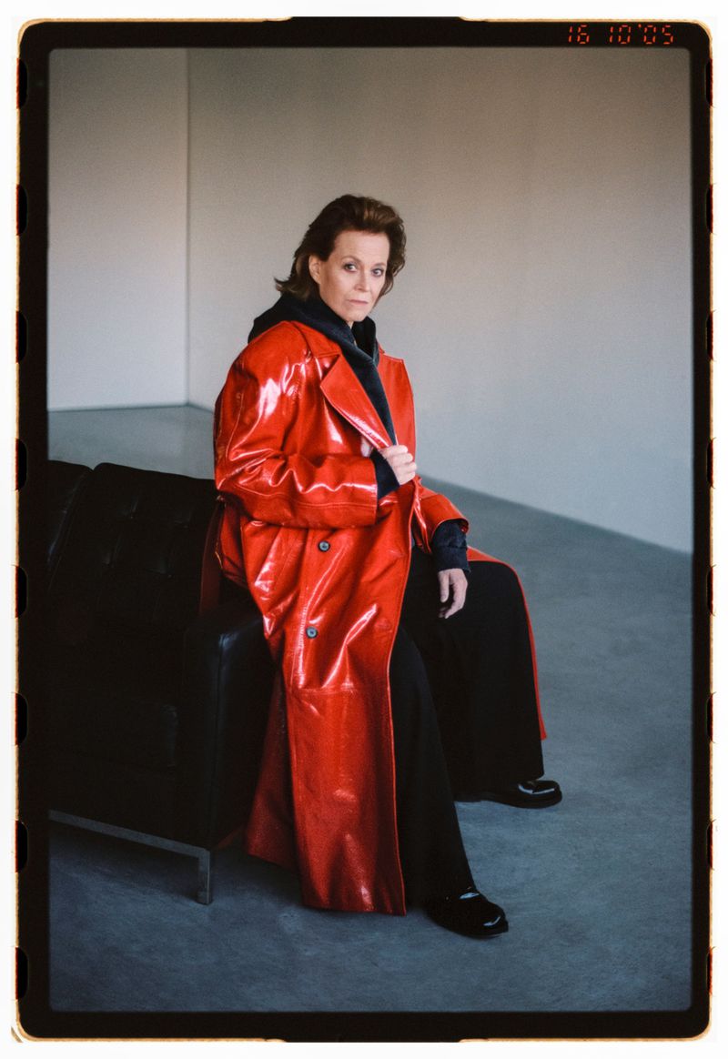 Stylish woman in a red trench coat sits on a black couch against a neutral wall, capturing a blend of fashion and portraiture.