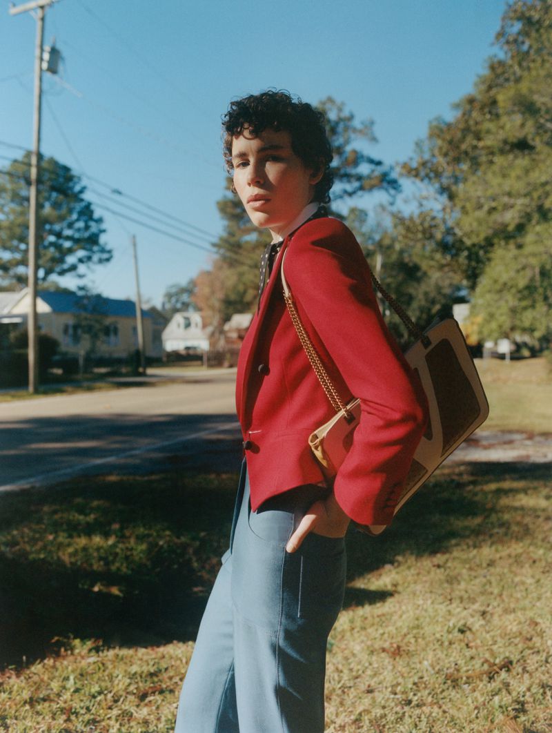 Young adult in red blazer with vintage bag stands on grassy lawn in front of suburban homes under a clear blue sky. Fashion editorial.
