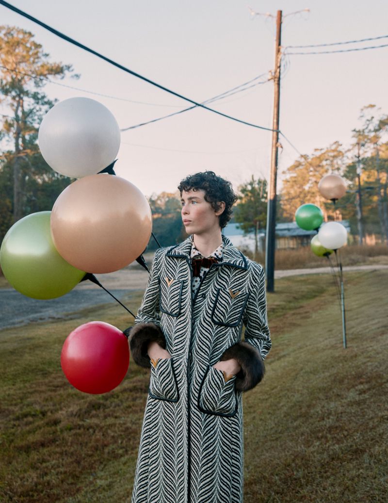 A woman in a patterned coat stands in a grassy field with colorful balloons against a backdrop of trees and a blue sky.