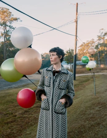 A woman in a patterned coat stands in a grassy field with colorful balloons against a backdrop of trees and a blue sky.