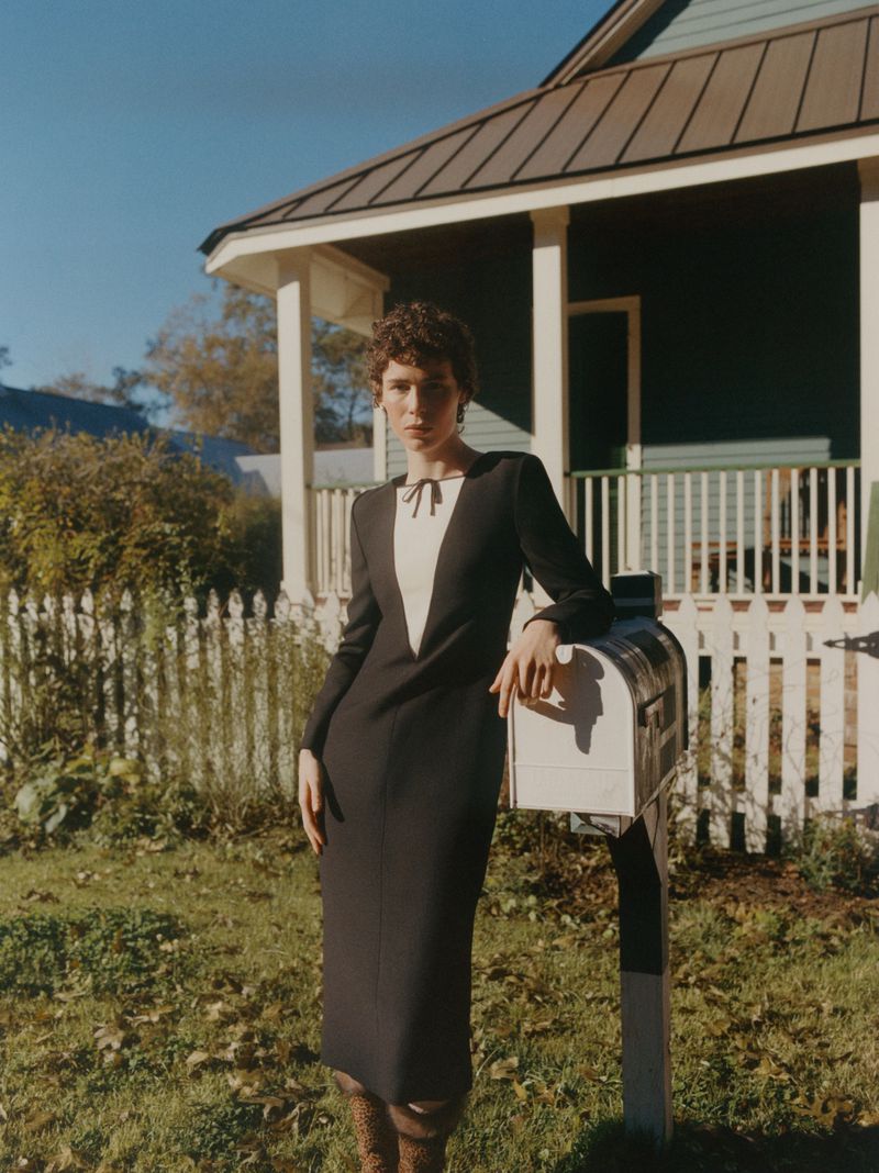 Woman in elegant black dress poses by a mailbox and vintage house on a sunny day, creating a retro-inspired fashion image.