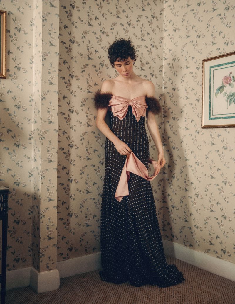 Person poses in a dark dress with a pink bow and fur trim against a floral wallpaper backdrop in an indoor environment.