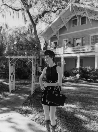 Young woman in 1960s dress poses on sidewalk with purse, in front of American house and garden on a sunny day. Black and white.