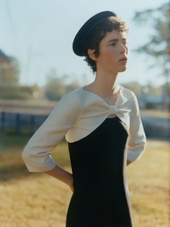 Model Jesi Evans wearing a black beret and an ivory bow-front shrug over a black dress against a soft outdoor background.
