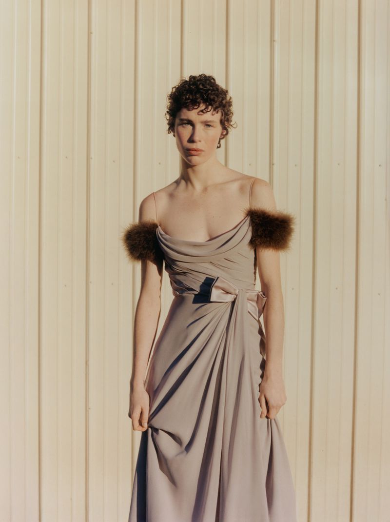 A person with curly hair poses in a flowing gown with fur accents against a neutral wall. The image has soft lighting and a studio feel.