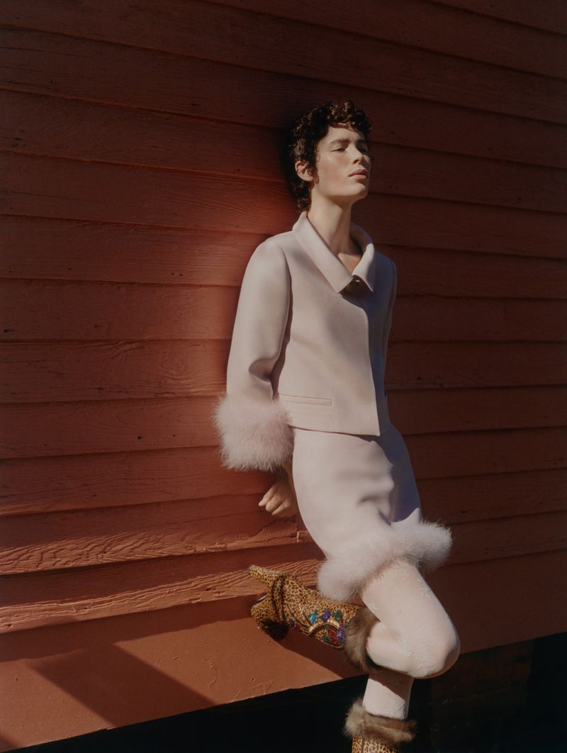 A young woman with short hair in a light-colored fur-trimmed suit and boots poses in front of a red wall in a fashionable pose.