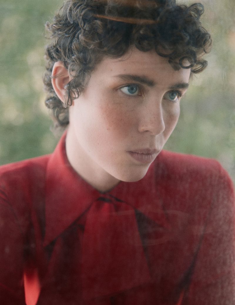 Close-up portrait of a young adult with curly brown hair and blue eyes, wearing a red blouse, looking down with a blurred green background.