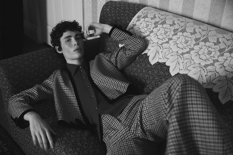 Black and white studio shot of a young adult male with curly hair laying on a patterned couch wearing a houndstooth suit and collared shirt.