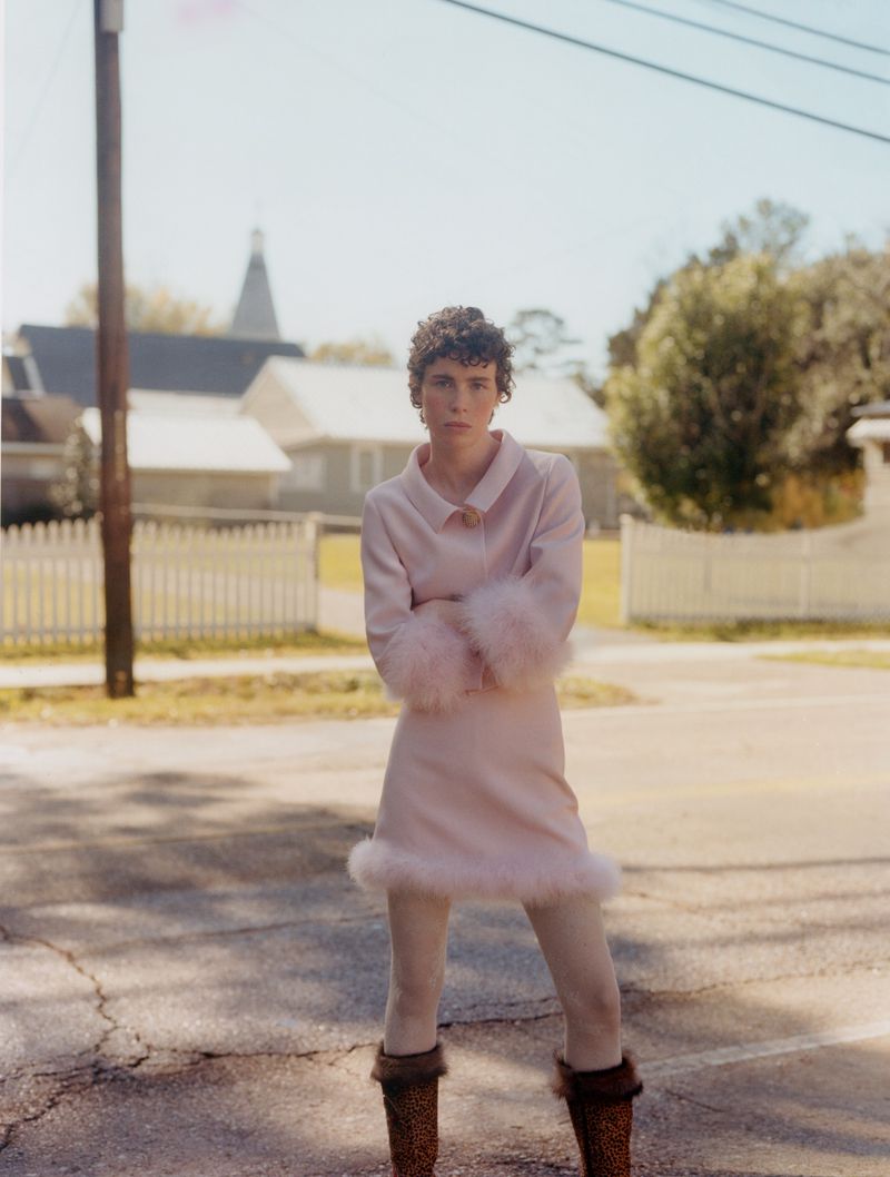 Woman in pink dress with fur cuffs and hem standing on an asphalt street with houses and trees in the background on a sunny day.