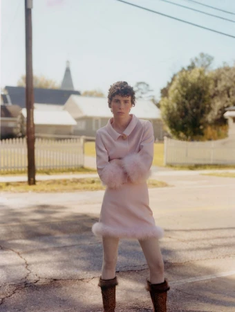 Woman in pink dress with fur cuffs and hem standing on an asphalt street with houses and trees in the background on a sunny day.