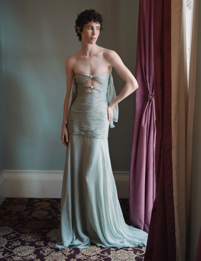 A woman in a light blue strapless gown stands in a room with a vintage carpet and mauve curtains, gazing toward a window with natural light.