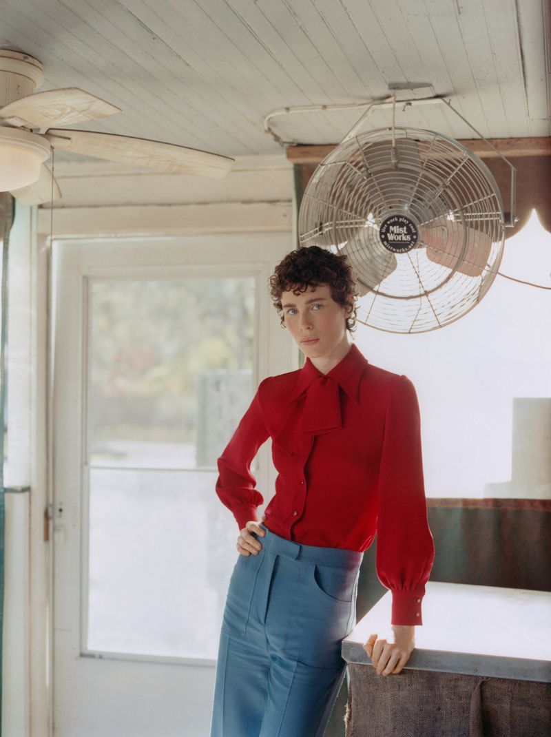 A person with short, curly brown hair poses indoors wearing a red blouse with a tie and light blue pants against a white wall.