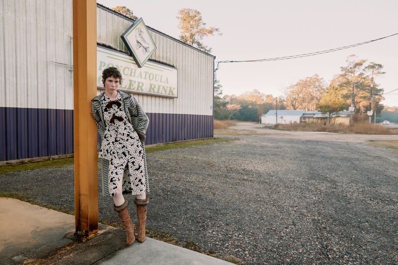 Woman in patterned dress and boots leans against building at Ponchatoula Roller Rink on overcast day in rural setting near trees.