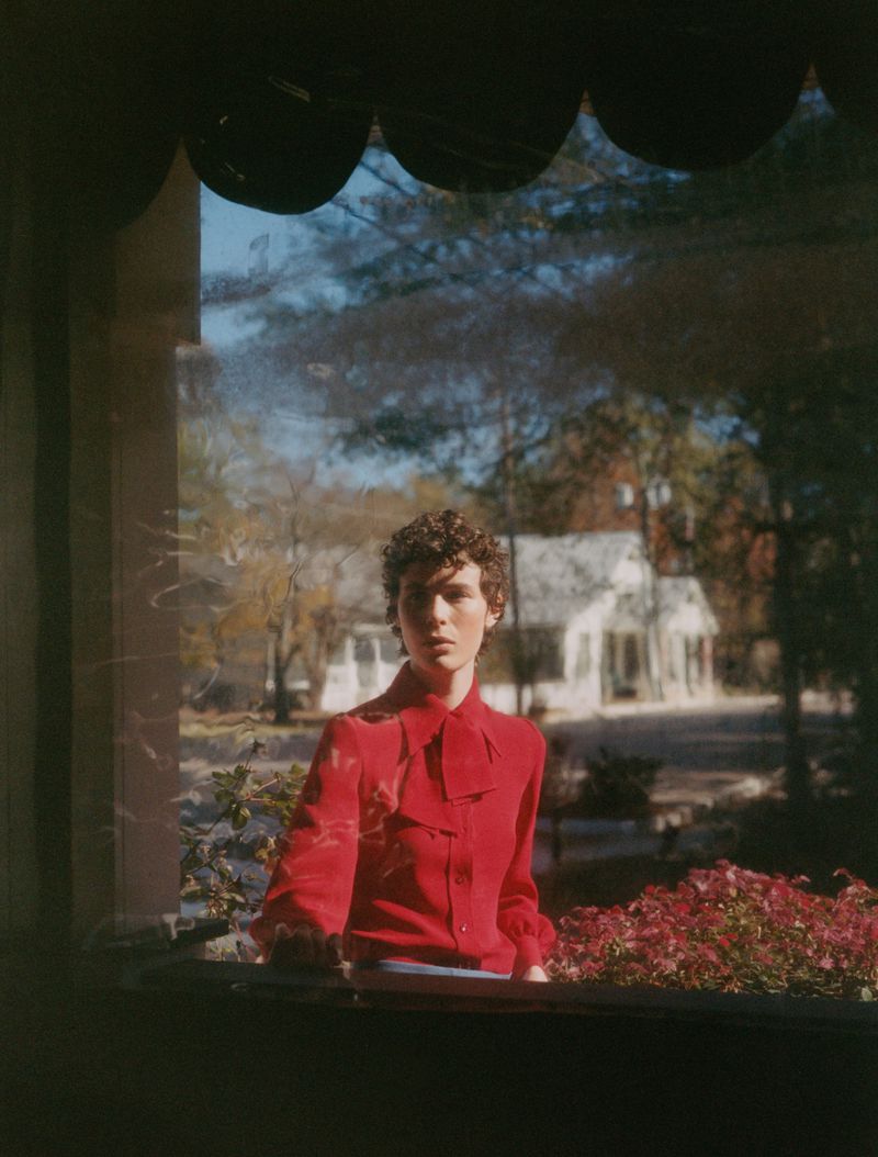 Portrait of a young adult with curly hair wearing a red top, photographed through a window with a view of a house and greenery.
