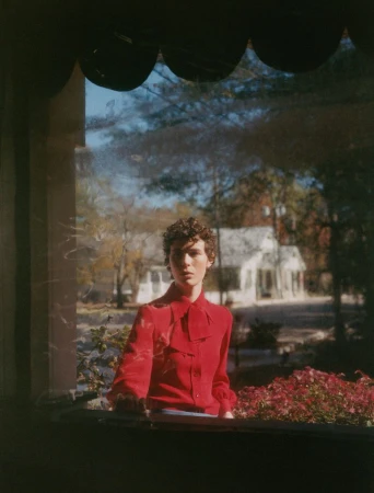 Portrait of a young adult with curly hair wearing a red top, photographed through a window with a view of a house and greenery.