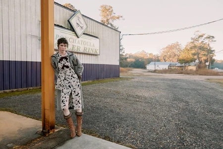 Model Jesi Evans leans against a pillar in a patterned coat and dress outside a rural roller rink.