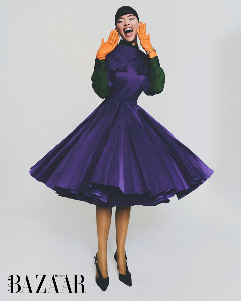 Fashion editorial shot of a woman in a purple dress, green top, and orange gloves, posing dynamically against a plain background with Harper's Bazaar logo.