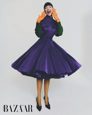 Fashion editorial shot of a woman in a purple dress, green top, and orange gloves, posing dynamically against a plain background with Harper's Bazaar logo.