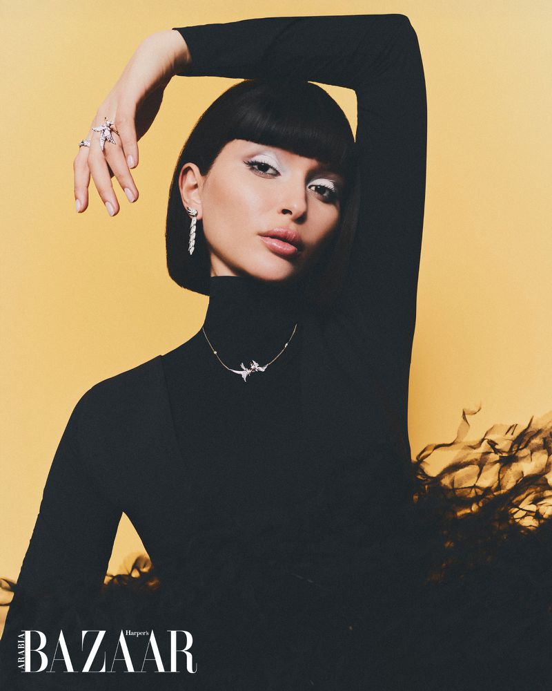 Portrait of a fashionable young adult woman wearing black turtleneck, white eye makeup, and jewelry against a yellow studio backdrop.