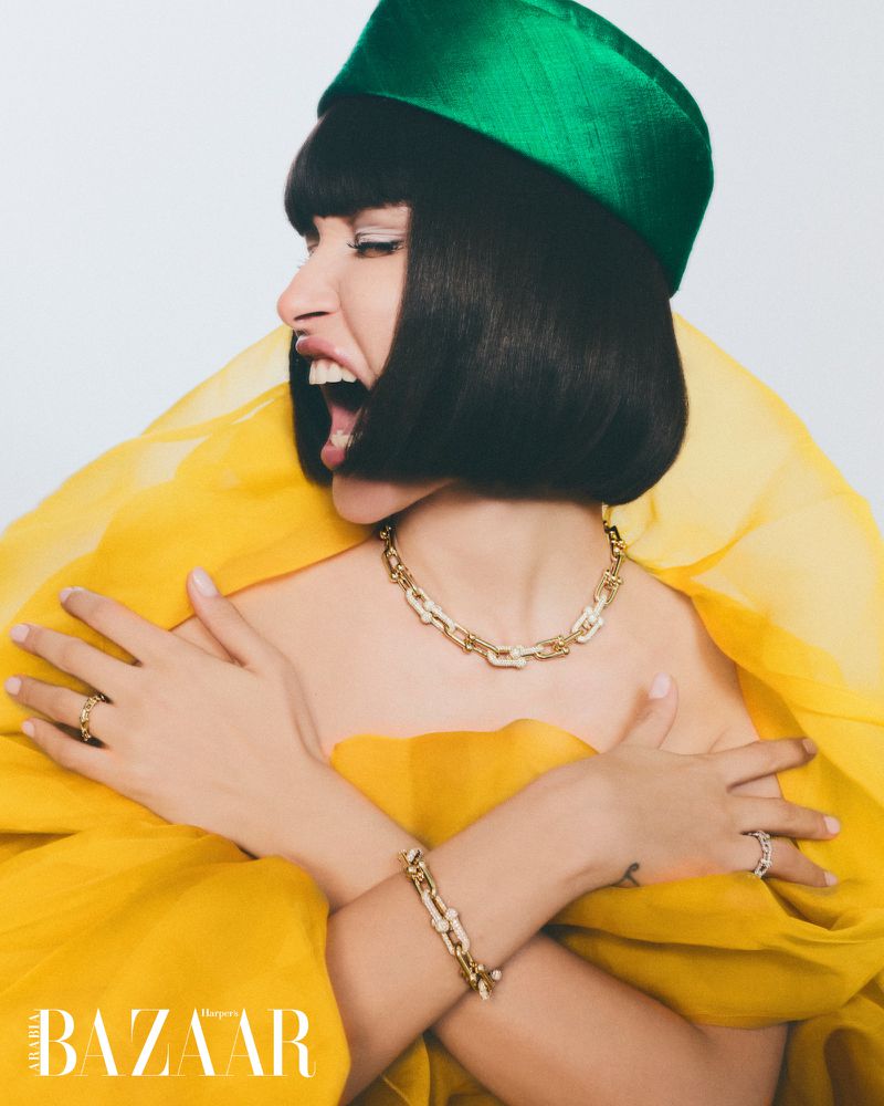 Profile shot of an adult woman with dark hair wearing a green hat, gold jewelry, and a yellow dress against a white backdrop.