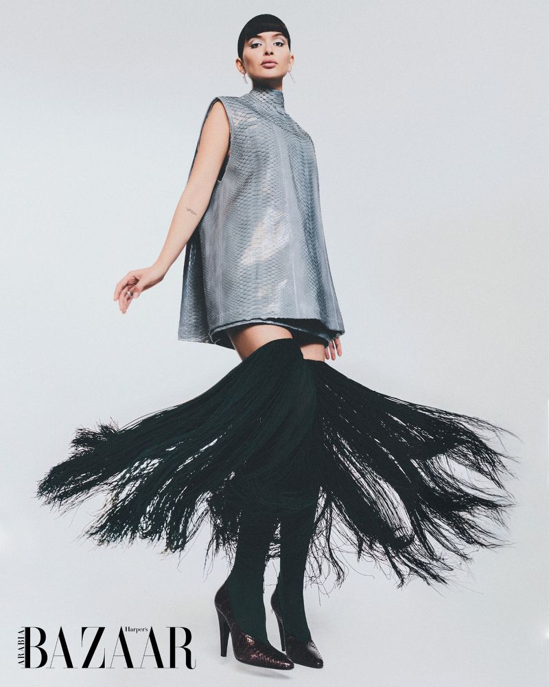 Fashionable woman posing in a shiny silver dress with black fringe thigh-high boots against a white background for Harper's Bazaar Arabia.