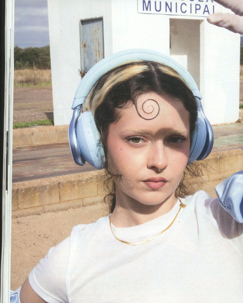 Close-up portrait of a young woman with spiral hair art wearing blue headphones, outdoors, with a white building in the background.
