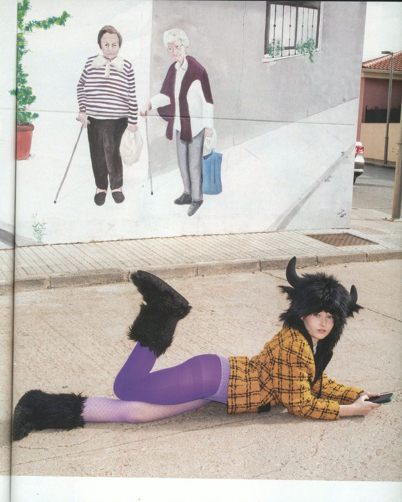 Young woman in costume lying on pavement in front of mural art featuring two elderly women and greenery; sunny day with natural light.