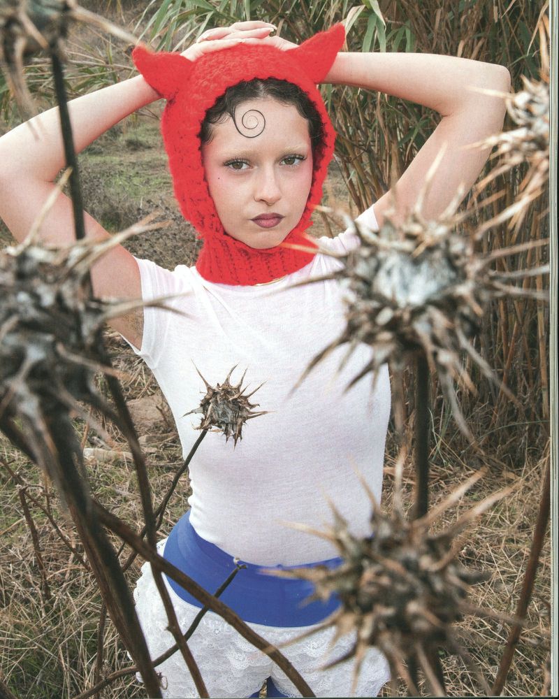 A young adult female in a red knit devil-eared cap poses outdoors among dry vegetation and spiky weeds. She wears a white t-shirt and shorts.