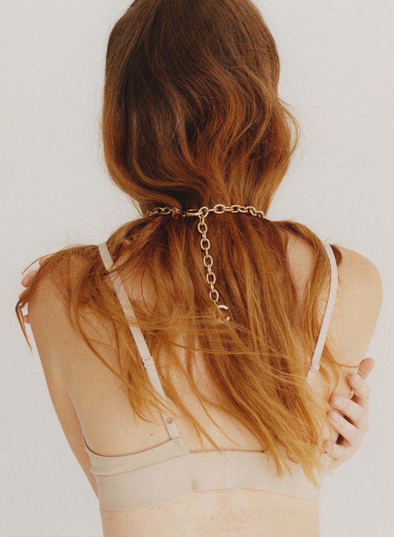 Back view of an adult female with long auburn hair wearing a beige bra and a gold chain necklace, against a white backdrop.