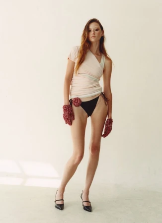 Fashion shot of a slender, red-haired woman wearing a cream-colored top, black bottoms, beaded mitts, and black high heels in a studio.