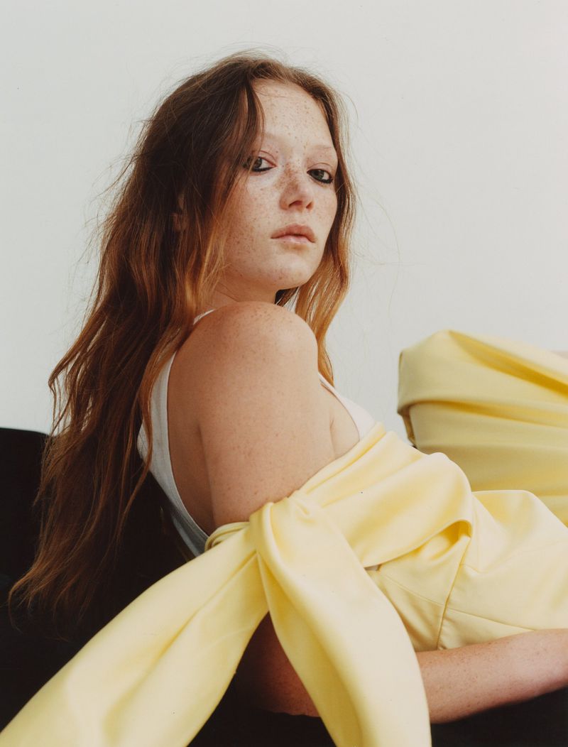 Red-haired young woman with freckles in a white top and yellow clothing sits indoors against a neutral wall in a portrait.