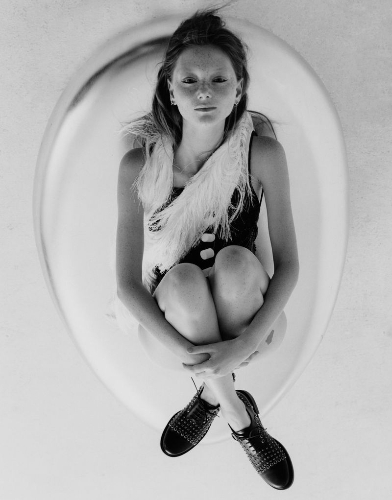 Top-down studio shot of a woman in a bodysuit and feather boa, seated inside a reflective dome, legs crossed, fair skin, dark shoes.